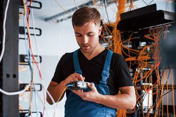 Wall Mural - Young man in uniform with measuring device works with internet equipment and wires in server room