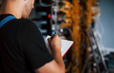 Wall Mural - Young man in uniform with notepad in hands works with internet equipment and wires in server room