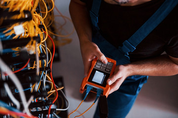 Wall Mural - Top view of young man in uniform with measuring device that works with internet equipment and wires in server room