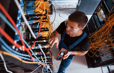 Wall Mural - Top view of young man in uniform with measuring device that works with internet equipment and wires in server room