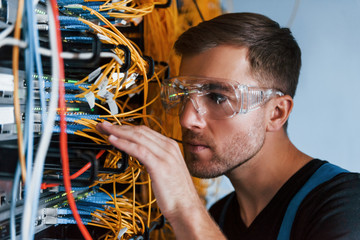 Wall Mural - Young man in protective glasses works with internet equipment and wires in server room