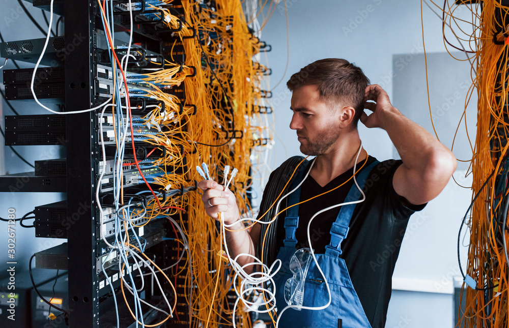 Young man in uniform feels confused and looking for a solution with internet equipment and wires in server room - obrazy, fototapety, plakaty 