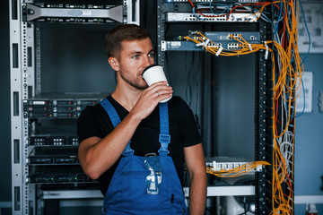 Wall Mural - Young man in uniform drinking coffee against internet equipment and wires in server room