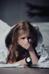 Wall Mural - scared child holding flashlight while hiding under blanket on black background with shadows