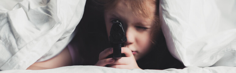 Wall Mural - panoramic shot of scared child holding gun while hiding under blanket