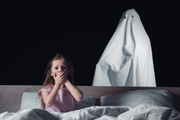 Wall Mural - frightened child sitting in bed and showing hush sign while white ghost standing behind bed isolated on black