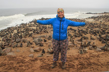 Young Caucasian Red-haired Happy Girl Traveler Backpacker In A Blue Down Jacket With Spread Out Arms Against The Background Of A Huge Colony Of Fur Seals In Cape Kros In Namibia, Africa.