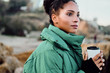 Side view of beautiful African American girl in down jacket with cup to go dreamily looking away on seaside