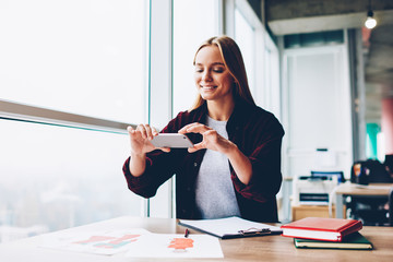 Canvas Print - Cheerful blonde female designer making photos of fashion sketches on modern smartphone sitting at desktop in office.Positive young woman taking pictures on mobile phone working in stylish studio