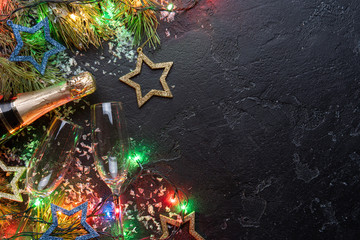 Image of branches of fir, Christmas decorations blue and gold toys, burning garlands, champagne bottles, two wine glass on black background