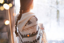 Thoughtful Young Brunette Woman Wearing Nordic Print Poncho Looking Through The Window, Blurry Winter Woods Snow Landscape Outside