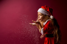 Little Blonde Girl In A Santa Suit Blows Snow Off Her Hands. Red Background Isolate. Space For Text