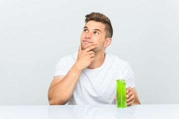 Wall Mural - Young handsome man holding a aloe vera bottle looking sideways with doubtful and skeptical expression.