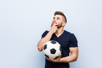 Wall Mural - Young caucasian man holding a soccer ball looking sideways with doubtful and skeptical expression.