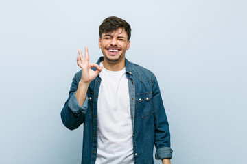 Young hispanic cool man winks an eye and holds an okay gesture with hand.