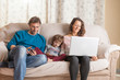 © selugallego - Child, a woman and a man sitting on a sofa watching a laptop and a smart phone.