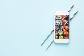 Mobile phone with sushi on screen and chopsticks on color background