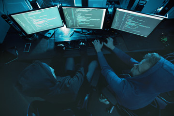 Rear view of computer hackers sitting in front of computer monitors and typing they breaking the system and working in team