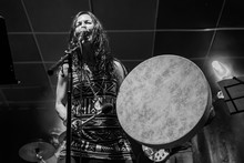 A Female Musician Is Viewed From A Low Angle As She Sings And Playing Native American Traditional Drum, Blurry Band Members Are Seen Performing In Background