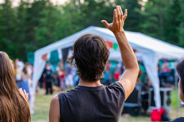 Poster - A young caucasian man is seen from behind with one arm raised in air during a shamanic exercises, diverse people enjoy spiritual gathering in the background