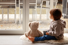 Cute Little Toddler Boy Sitting At Window With Cup Of Warm Tea With His Soft Teddy Bear Toy Looking Out Thoughtfully At Chilly Autumn Weather. Cozy Home. Fall Melancholy Concept. Seasonal Mood