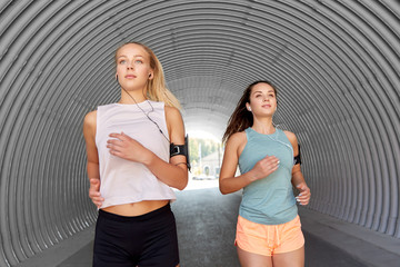 Wall Mural - fitness, sport and healthy lifestyle concept - young women or female friends with earphones wearing armbands with smartphones and running outdoors