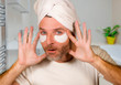 © TheVisualsYouNeed - happy and attractive camp gay man applying moisturizer eye patch facial product with head wrapped in towel looking playful bathroom mirror in proud feminine look
