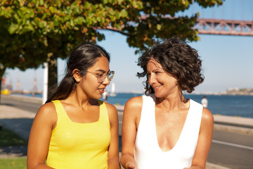 Wall Mural - Cheerful multiethnic women walking at riverside. Beautiful smiling female friends walking and talking near river. Friendship concept