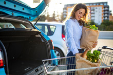 Caucasian Brunette Going Holding Paper Bags With Food Products. Young Woman Putting Package With Groceries And Vegetables Into Car Trunk. Attractive Caucasian Female Shopping In Mall Or Grocery Store