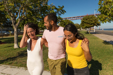 Wall Mural - Cheerful friends embracing in park. Group of happy multiethnic friends walking together and laughing at riverside. Friendship concept