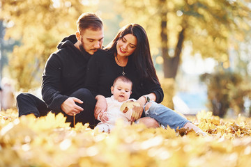 Wall Mural - Cheerful family sits on the ground and having fun together with their child in beautiful autumn park