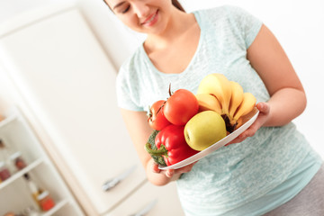 Canvas Print - Body Care. Chubby girl standing in kitchen with plate of fruits and vegetables close-up smiling happy blurred