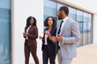 Positive multiethnic business colleagues walking near office building. Man and women wearing formal suits, going past outdoor wall, talking to each other. Corporate friends concept