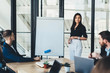 © BullRun - Portrait of confident business woman standing near flip chart for presentation and looking at camera, young male and female colleagues using white board with copy space area for your advertising