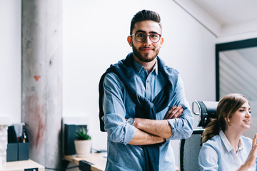 Half length portrait of positive successful man specialist looking at camera while standing with crossed arms in office interior while colleagues talking on background, job and occupation