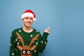 Happy young man in a Christmas sweater and hat isolated on a blue background, points his finger away at copy space, looks into the camera and smiles. Isolated.