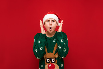 Portrait of surprised young man looking into camera, screaming with shock with raised hands, isolated on red background, wearing Christmas hat and sweater. Copy space