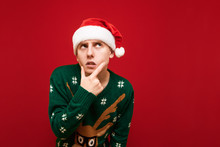Close Up Portrait Of A Funny Pensive Guy In Christmas Clothes, Wearing A Santa Hat And A Green Warm Sweater, Looking Away And Thinking. Funny Guy With A Christmas Mood Thinks With A Serious Face.