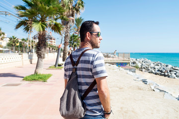 Man with shoulder bag standing on promenade looking to horizon in a sunny day