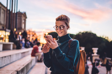 Wall Mural - Positive young hipster girl typing text message using 4g wireless for networking and online communication, happy female tourist in spectacles blogging creating publication about tourism and trip