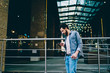 © BullRun - Side view of casual dressed hipster guy strolling in urban setting near business building and reading received notification on smartphone.Young man chatting in social networks on cellular on street