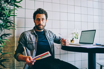 Wall Mural - Portrait of smart bearded young man looking at camera while holding paper report in hand and working on accounting data at laptop computer.Intelligent hipster guy in casual wear sitting at table