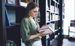 © BullRun - Student thoughtfully reading book leaning on bookshelves..Side view of smart young casual brunette in glasses and with long hair immersed in reading book standing near bookshelves in light library