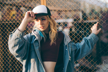 Young attractive woman in jeans jacket, shorts, red top and trucker hat posing over metal fence and graffiti wall over background in a city