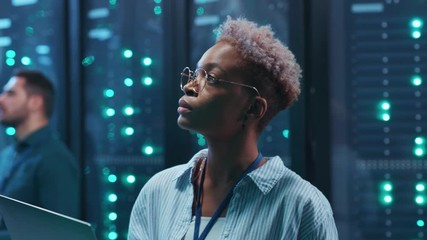 Beautiful african woman IT technician using laptop diagnosing hardware security system on network servers working in rack corridor. Close-up. Big data center.
