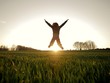 Joyful and happy girl is jumping with open arms on a green field at sunset with the sun in the background