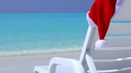 Wall Mural - Santa Claus helper Hat on sunbed near calm beach with azure sea water and white sand. Christmas vacation on island