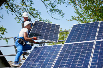two workers installing heavy solar photo voltaic panel on tall steel platform on green tree and blue