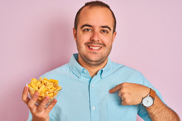 Canvas Print - Young man holding bowl with macaroni pasta standing over isolated pink background with surprise face pointing finger to himself