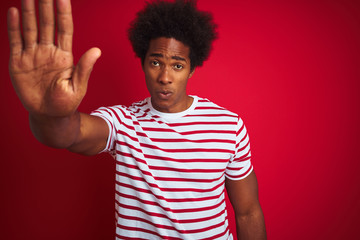 Canvas Print - Young african american man with afro hair wearing striped t-shirt over isolated red background doing stop sing with palm of the hand. Warning expression with negative and serious gesture on the face.